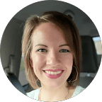 A woman with short brown hair and a light top smiles at the camera inside a car.