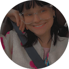 A person with glasses and dark hair smiles while sitting in a car, wearing a light-colored jacket and a seatbelt.