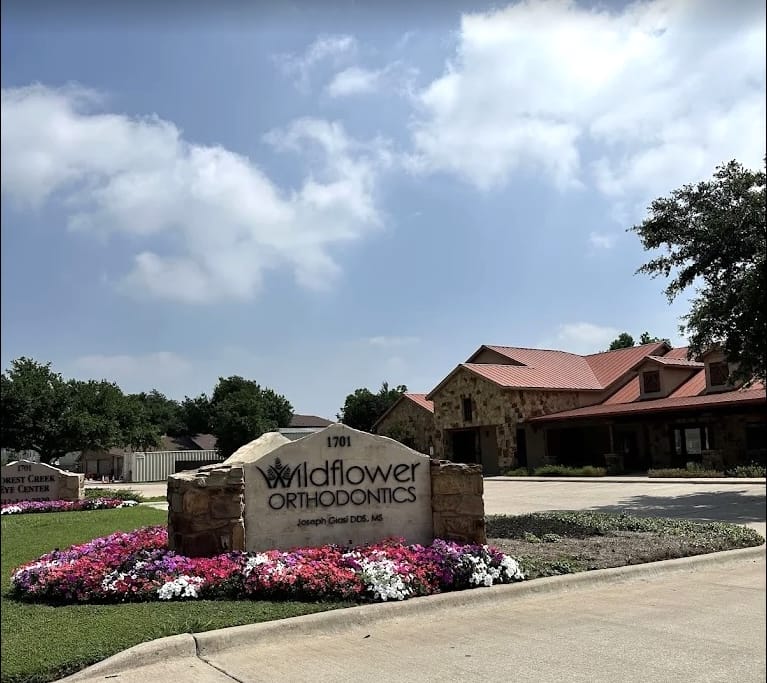 About the stone sign for Wildflower Orthodontics: it's surrounded by flower beds in front of a building with a red roof, all set beneath a partly cloudy sky.