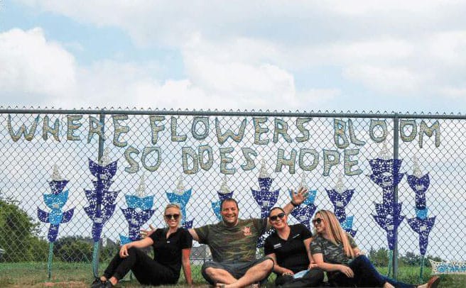 Four people sit smiling in front of a chain-link fence decorated with the words "WHERE FLOWERS BLOOM SO DOES HOPE," flower designs, and a sense of community.