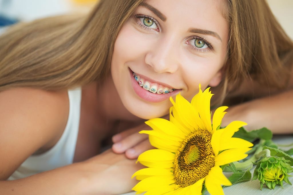 Young woman with long hair and braces smiles while lying down, holding a large yellow sunflower close to her face.