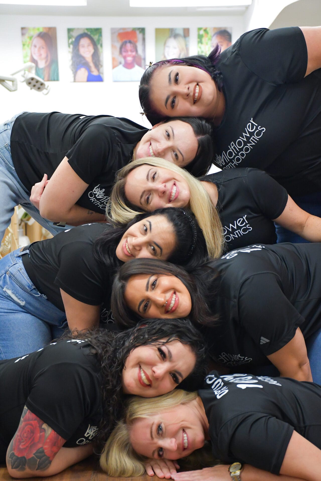 Seven women from our team, wearing matching black shirts, are stacked in a pyramid formation, smiling at the camera in an indoor setting with portraits visible on the wall behind them.