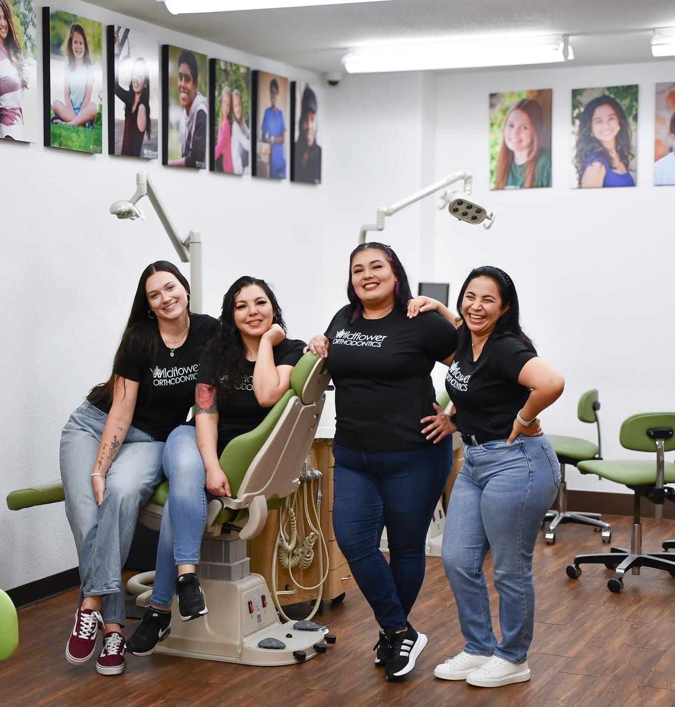 Four women from our team wear matching shirts as they pose and smile in a dental office, standing and sitting around a dental chair. Portraits are displayed on the wall behind them.