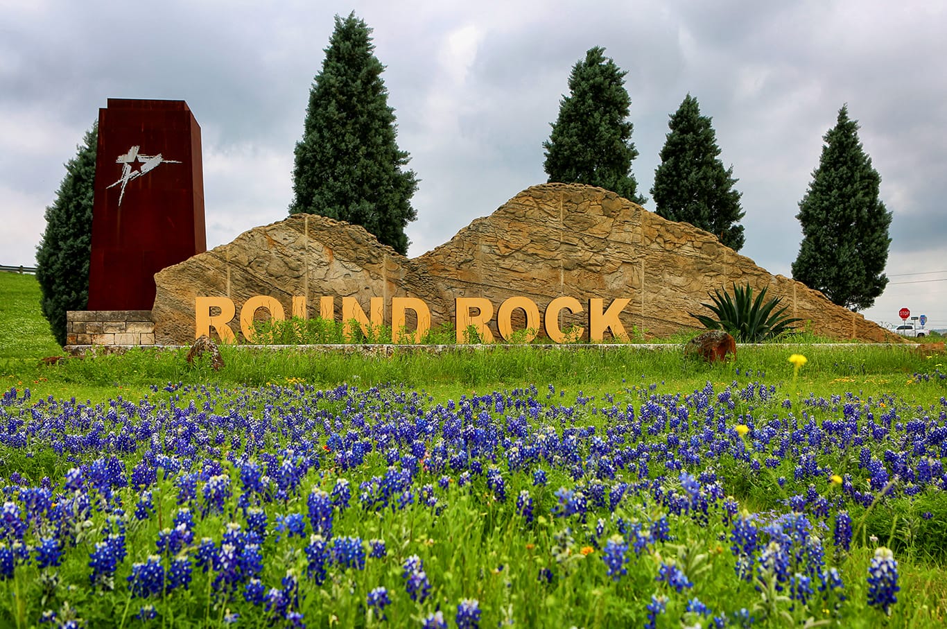 Stone Round Rock city sign with large yellow letters, red monument, evergreen trees, and blooming bluebonnet flowers in the foreground—showcasing community support under a cloudy sky.