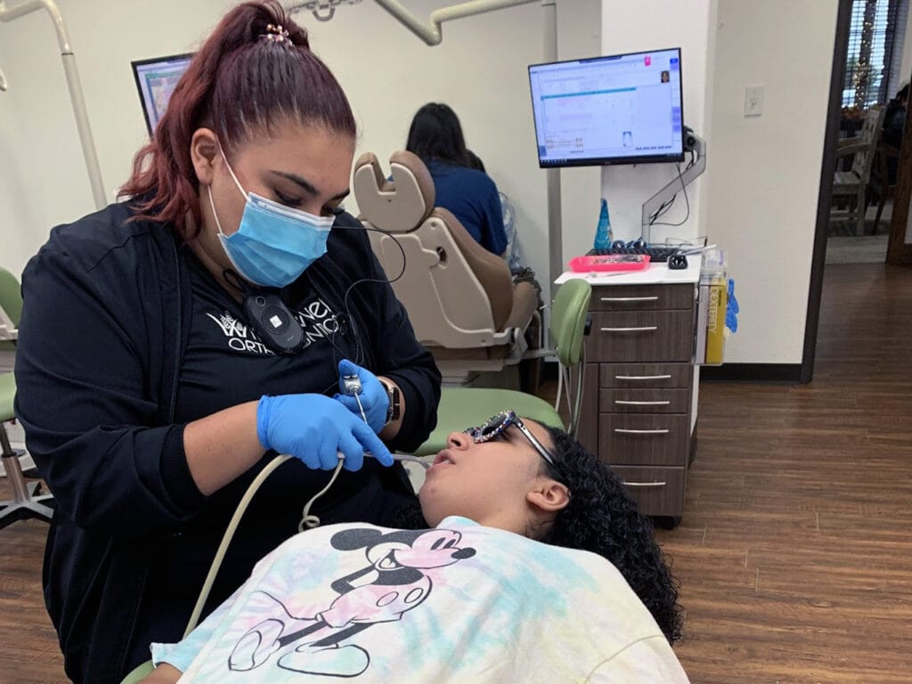 A dental professional in a mask and gloves works on a patient lying in a dental chair, with dental equipment and a monitor visible in the background.
