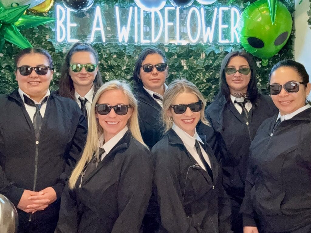 Six women in black suits and sunglasses stand in front of a green foliage wall with a neon sign that says "BE A WILDFLOWER" and large green alien decorations.