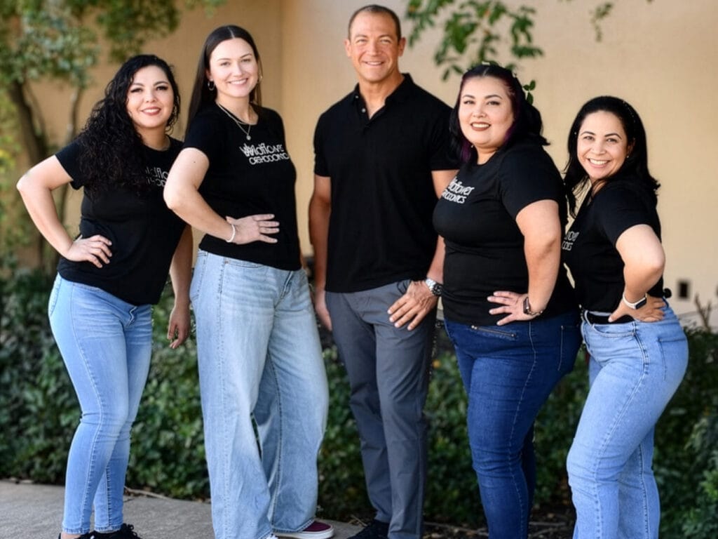 Five people, four women and one man, stand side by side outdoors, all wearing black shirts and smiling at the camera. Shrubbery and a beige wall are in the background.