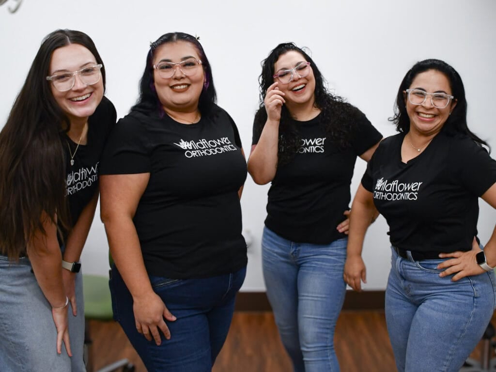 Four women wearing glasses, black "Wildflower Orthodontics" shirts, and jeans stand together smiling in a room with white walls and wood flooring.