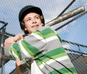 A teenage boy with braces, wearing a helmet and striped green shirt, holds a baseball bat, standing in a batting stance at a fenced baseball field.